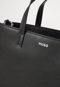 Svart lær tote-veske med teksturert overflate, to håndtak og en sølvfarget "HUGO"-logo. Åpen toppdesign med romslig interiør.