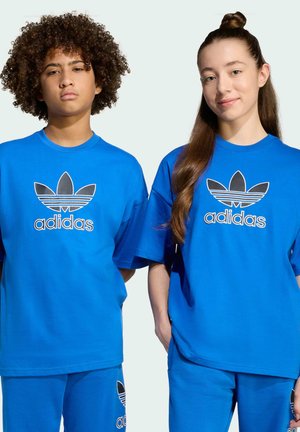 Dos adolescentes con camisetas y pantalones Adidas a juego de color azul brillante, de pie uno al lado del otro contra un fondo blanco liso.