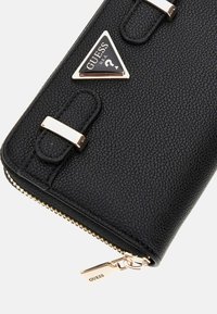 Portafoglio in pelle nera con texture, dotato di hardware dorato, con un'etichetta logo triangolare e chiusura a zip. Forma rettangolare compatta.