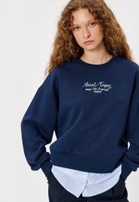 Sudadera azul marino con el texto bordado "Saint Tropez Sailing Club", puños acanalados y un corte relajado sobre una camisa de rayas azul claro.