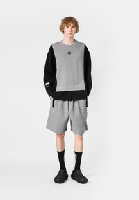 Sudadera gris con mangas negras, logo en el pecho, combinada con pantalones cortos grises con detalle de cadena. Calzado y calcetines negros completan el conjunto.