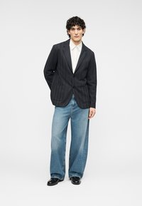 Μαύρο υφασμάτινο σακάκι με δύο μπροστινές τσέπες, συνδυασμένο με ανοιχτό μπλε τζιν παντελόνι φαρδιάς γραμμής και μαύρα παπούτσια. Το μοντέλο στέκεται σε ουδέτερο φόντο.