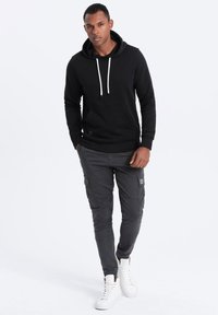 Sudadera negra con capucha y cordones blancos, combinada con pantalones cargo grises que tienen bolsillos laterales y un acabado texturizado. Zapatillas blancas completan el look.