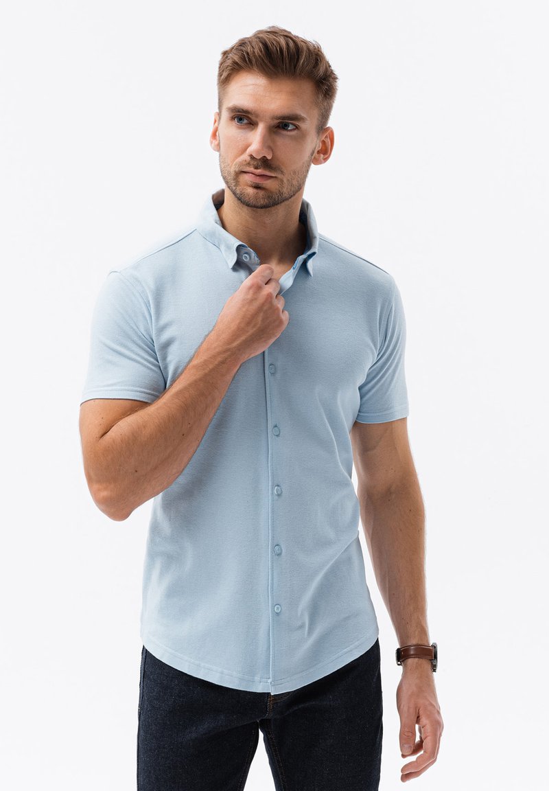 Lichtblauwe polo met korte mouwen, voorzien van een klassieke kraag, knoopsluiting aan de voorkant en een gestructureerde stof. G gedragen met donkere jeans.