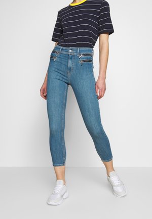 Jeans ajustados de denim azul claro con bolsillos delanteros con cremallera. Combinados con una camiseta de rayas en azul marino y zapatillas blancas.