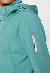 Jachetă de exterior teal cu un buzunar cu fermoar pe mâneca stângă. Prezintă o textură netedă și o glugă ajustabilă cu șnururi.