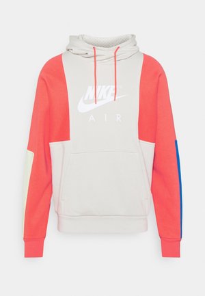 Bluza z kapturem Nike Air z jasnobeżowym tułowiem, koralowymi rękawami, niebiesko-kremowymi łatami na łokciach, przednią kieszenią i koralowymi ściągaczami na kapturze.