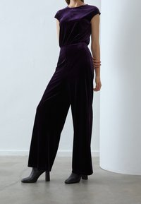 Samet jumpsuit i djup lila med cinchad midja och vida ben; kort ärm och slät textur; bärs med svarta klackade ankelstövlar.