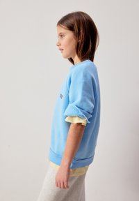 Jeune fille en sweat-shirt bleu clair aux manches retroussées, chemise beige en dessous, et pantalon de couleur claire, de profil sur un fond uni.