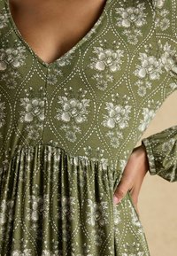 Zelené šaty s výstrihom do V, dlhými rukávmi, so vzorom kvetov v bielej farbe, textúrovanou látkou a s jemným nariasením v oblasti pásu.