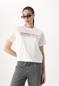 Bílé bavlněné tričko s crop střihem a hravým nápisem "HOWDY" v hnědém písmu. Vytvořeno v kombinaci se šedými džíny.