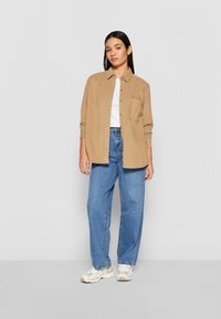 Chemise boutonnée brun clair avec une poche, portée sur un haut blanc, associée à un jean bleu ample et des baskets beiges. Texture du tissu lisse.