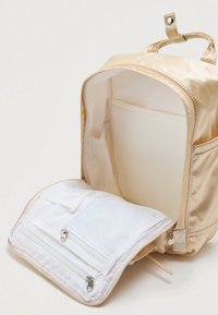 Μπεζ σακίδιο νάιλον με λεία υφή και στρογγυλή μορφή. Το εσωτερικό διαθέτει λευκό τμήμα με φερμουάρ και τσέπη από δίχτυ για οργάνωση.