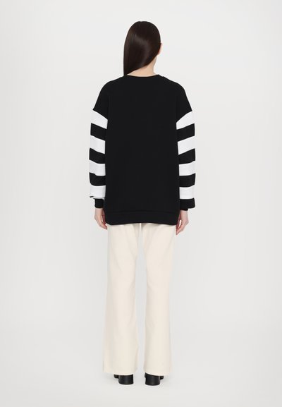 Black sweatshirt with wide white stripes on sleeves, relaxed fit, paired with flared cream trousers and black ankle boots. Smooth texture.