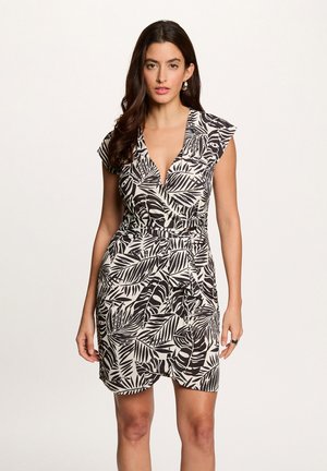 Donna che indossa un abito avvolgente a foglie bianche e nere con scollo a V e maniche corte, in piedi davanti a uno sfondo chiaro e semplice.