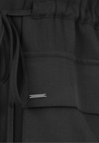 Indumento in tessuto nero con vita a coulisse, superficie strutturata e una piccola etichetta argento con testo in rilievo.