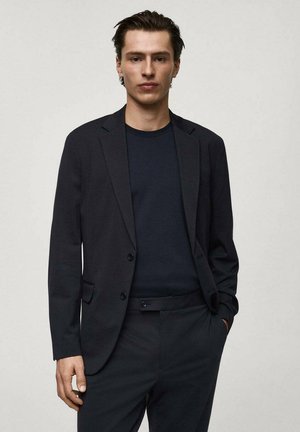 Black tailored blazer with a notched lapel, single-button closure, and front pockets, worn over a dark shirt and matching trousers.