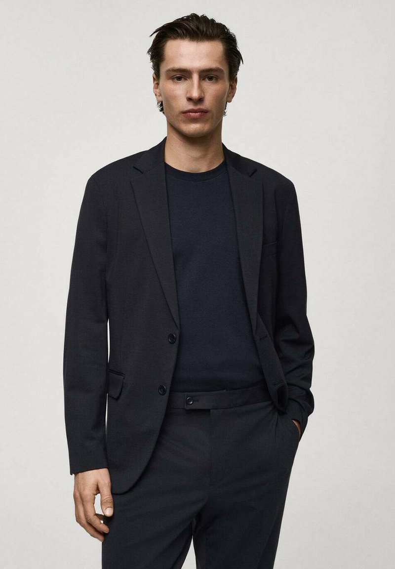Blazer nero su misura con revers a punta, chiusura con un bottone e tasche frontali, indossato sopra una camicia scura e pantaloni coordinati.