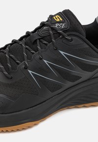 Baskets de sport noires avec un dessus en maille texturée, lacets élastiques, accents réfléchissants et une semelle en caoutchouc épaisse avec une bande de roulement de couleur gomme.