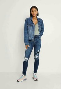 Casaco de ganga, blusa verde claro, jeans azuis desgastados com rasgos. Ténis multicoloridos claros com um acabamento refletor. Design simples.