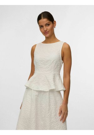 Ženska z lasmi zvezanimi nazaj, oblečena v brezrokavnik z belim teksturiranim peplum zgornjim delom in ujemajočo se krilo, na svetli enobarvni podlagi.