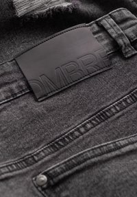 Primer plano de unos pantalones de mezclilla gris oscuro que muestran un parche de cuero cosido con el nombre de la marca parcialmente grabado y un lazo para el cinturón visible, así como la costura del bolsillo.