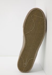 Semelle d'une chaussure beige avec un motif de bande de roulement en zigzag, appuyée contre un mur blanc et posée sur une surface blanche.