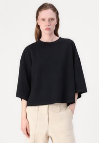 Μαύρη βαμβακερή oversized μπλούζα με κοντό μανίκι, στρογγυλή λαιμόκοψη και κοντό τελείωμα, συνδυασμένη με καφέ παντελόνι.