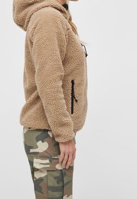 Giacca a felpa beige con cappuccio e superficie testurizzata, tasche laterali e cerniere nere. Indossata con pantaloni cargo mimetici.
