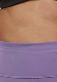 Leggings de sport violettes avec une texture lisse, dotées d'une large taille. Teint de peau visible au-dessus de la taille. Détails de couture soignés.