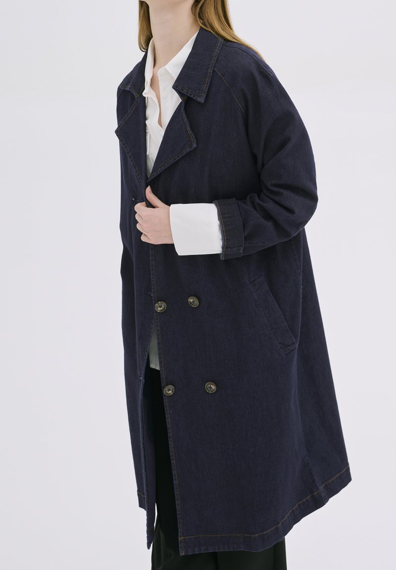 Mørk denimfrakke med dobbeltradet design, store revers, sidelommer og kontrastsyninger. Lang, løs silhuet.