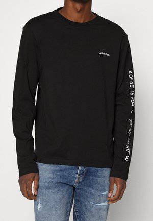 Homme portant un t-shirt noir à manches longues Calvin Klein avec des coordonnées blanches imprimées sur la manche droite et un jean bleu usé.