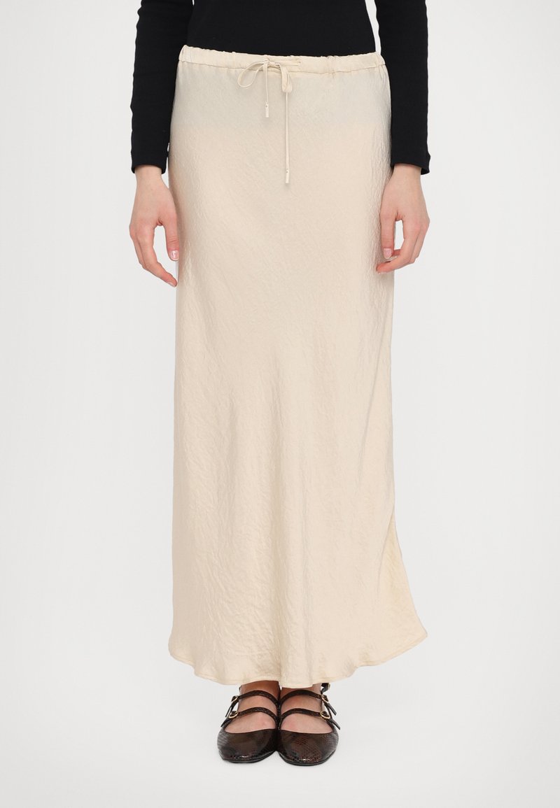 Jupe longue couleur crème avec une texture froissée, taille élastique à cordon de serrage, et une forme fluide. Associée à des chaussures plates noires.