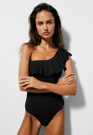 Kobieta z długimi ciemnymi włosami w czarnym, jednoczęściowym kostiumie kąpielowym na jedno ramię z falbanką, stojąca ze skrzyżowanymi ramionami na jednolitym tle.