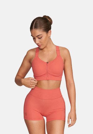 Kobieta regulująca koralowy sportowy stanik z przednim zamkiem błyskawicznym, w dopasowanych szortach z wysokim stanem, stojąca na tle jednolitego jasnego tła.
