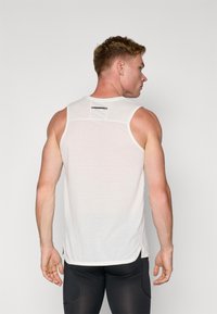 Biały sportowy top typu tank z wytrzymałą siateczkową plecą, okrągłym dekoltem i bocznymi rozcięciami. Na górnej części pleców znajduje się małe czarne logo.