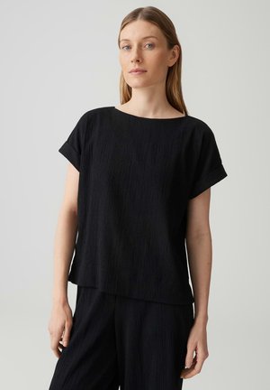 Frau mit glatten, hellbraunen Haaren, die ein locker sitzendes schwarzes Kurzarmoberteil und passende schwarze Hosen vor einfachem Hintergrund trägt.