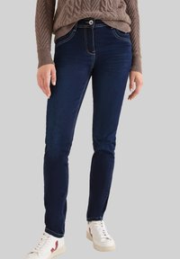 Jeans de denim azul oscuro con un ajuste slim, con costuras de contraste, una cintura media y cierre de botón. Combinados con zapatillas blancas.