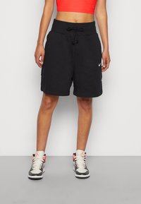 Person som bär svarta Nike shorts med dragsko, röd crop top och vita, svarta och röda högskaftade sneakers, står mot en grå bakgrund.