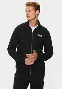 Uomo in giacca nera con zip completa e pantaloni coordinati, con il logo bianco di EA7 Emporio Armani, in piedi con le mani unite, mentre guarda verso destra.