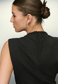 Naine, kelle juuksed on kokku pandud kuklasse, kannab ilma varrukateta musta topi ja rippuvaid hõbedasi kõrvarõngasid, kujutatud profiilis ühtlasel taustal.