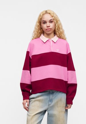 Jeune personne aux longs cheveux blonds bouclés portant un maillot de rugby rayé rose et bordeaux et un jean bleu clair, debout devant un fond blanc.