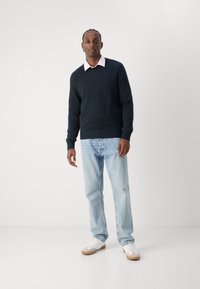 Maglione blu scuro con polsini e orlo a coste, indossato sopra una camicia bianca con colletto; jeans azzurri e sneakers bianche con dettagli beige.