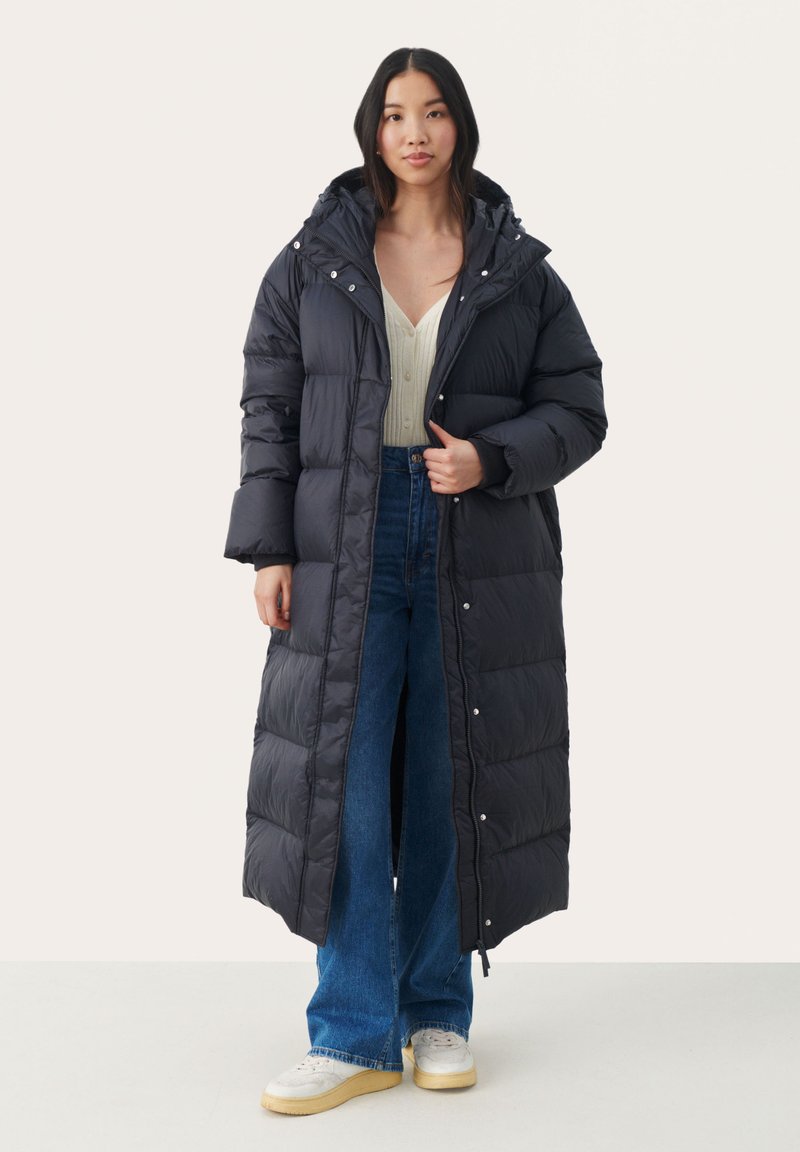 Lange zwarte pufferjas met een capuchon, geborduurde textuur, drukknoopsluiting en zijsplitten, gedragen over een gebreide top en wijde jeans.