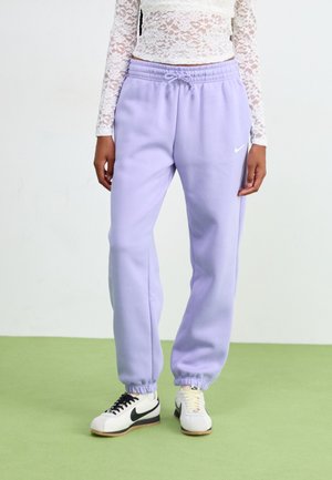 Kvinde iført lilla Nike joggingbukser med snøre i taljen, hvid blonder med lange ærmer og forskellige hvide sneakers med sorte swoosh.