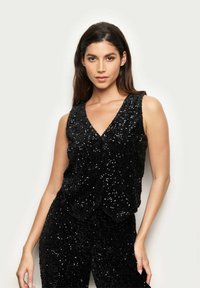 Czarny bezrękawnik z głębokim dekoltem w kształcie V, pokryty błyszczącymi cekinami, z teksturowanym materiałem i falbaniastym wykończeniem, które dodaje szczegółów.