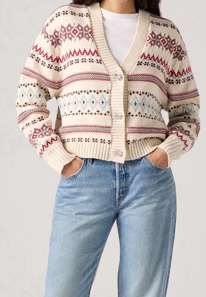 Personne portant un cardigan crème avec des motifs tricotés rouges et bleus, boutonné devant, associé à un jean bleu clair et les mains dans les poches.
