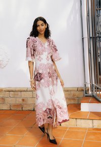 Femme en robe midi rose et blanche à motifs avec fente, talons noirs pointus, debout sur un sol carrelé orange près d'un mur en brique et blanc.