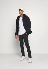 Fekete steppelt kapucnis dzseki fehér póló fölött; fekete, szűkített nadrág; fehér tornacipő; világos színű sapka. Minimalista kialakítás.