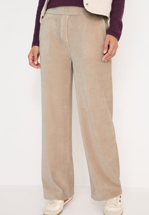 Beige wijde corduroy-broek gedragen met witte sneakers en een paarse langemouwen top, met ontspannen handen aan de zijkanten.
